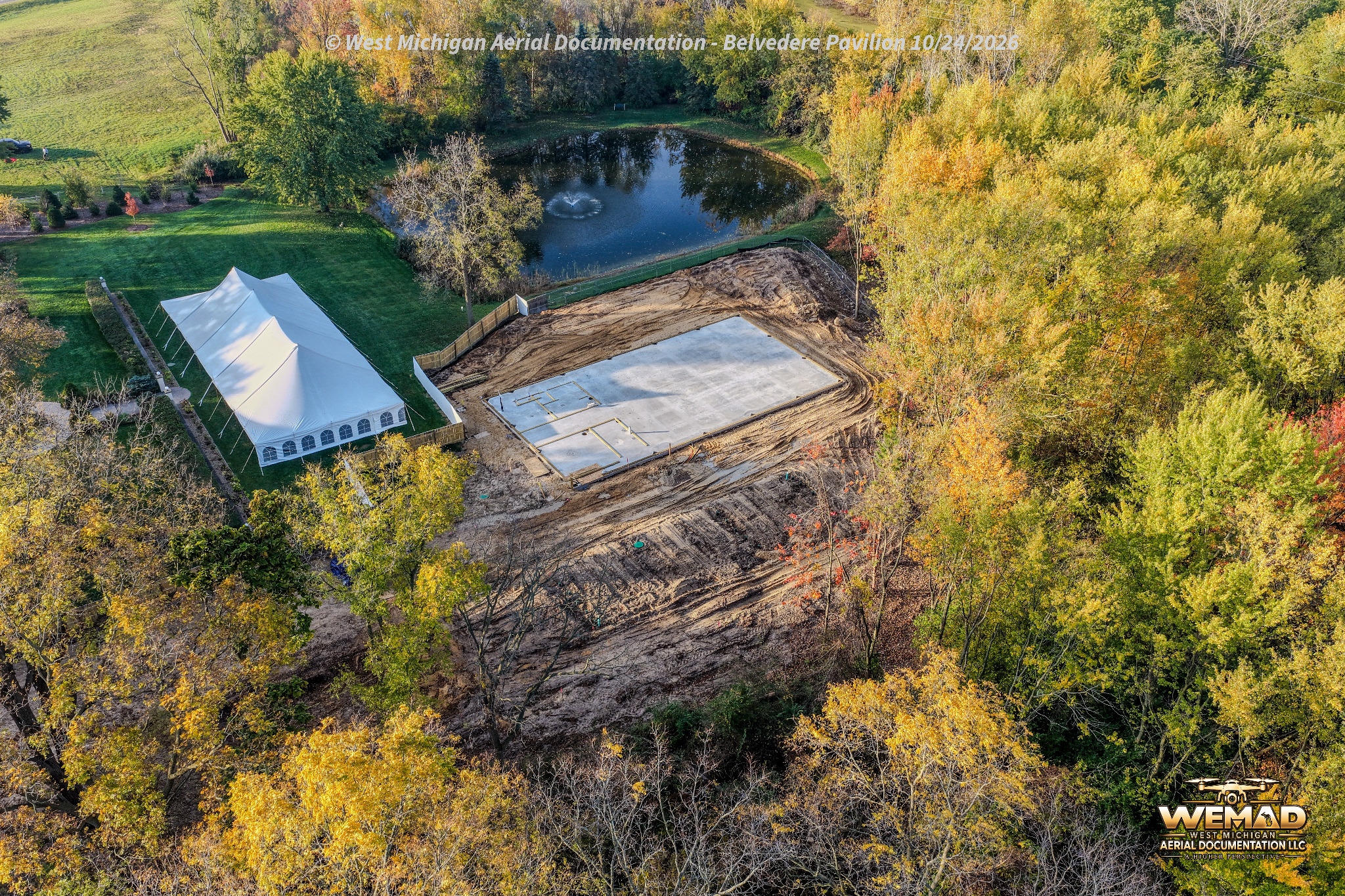 Reverse aerial view of The Belvedere pavilion concrete slab, site work, event tent, and pond, October 24, 2026